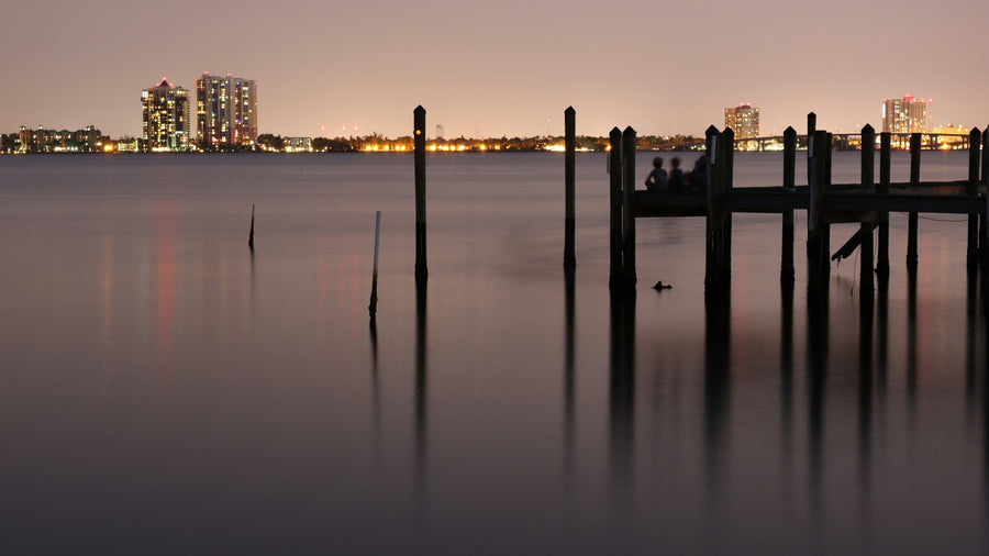 Downtown Fort Myers skyline lit up at dusk across the Caloosahatchee River, with old dock pilings in the water